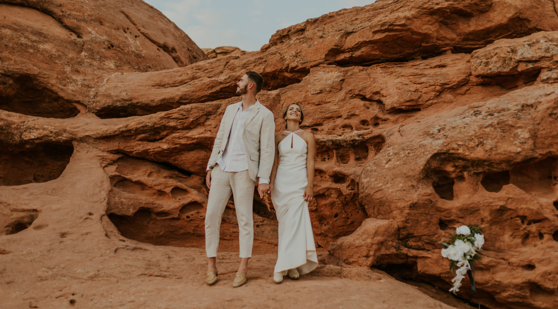 A groom and bride hold hands against red rock in Captiol Reef National Park in southern Utah. The bride's bouquet is sitting against a rock to the right of her.
